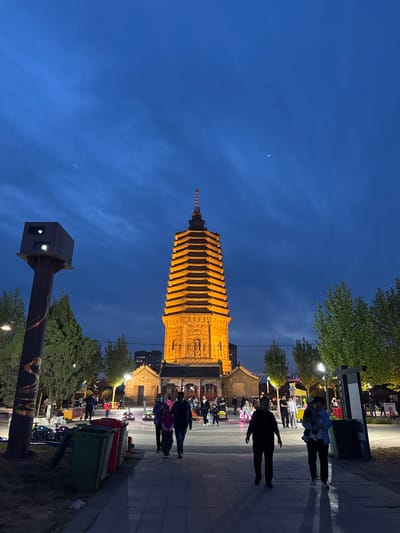 An outdoor nighttime scene featuring the Guangji Temple Pagoda in Jinzhou, China. The pagoda is the focal point, illuminated in a warm orange light, contrasted against a dark blue sky with a sliver of moon visible. In the foreground, a streetlamp with an attached black box stands beside a sidewalk. People are walking on the sidewalk near the base of the pagoda. A row of trash receptacles of various colors (green, red, and black) borders the sidewalk. Trees and a small building are also visible.