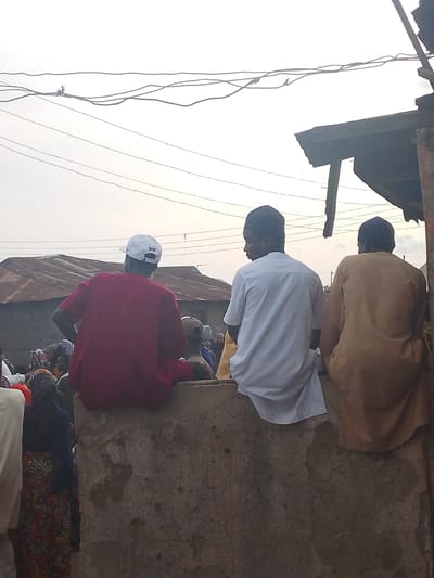 The image depicts a street scene in Tundun Wada South, Nigeria, with three men sitting on a low, rough concrete wall in the foreground, facing away from the camera. The man on the left wears a red t-shirt and a white baseball cap with the Nike logo. To his right, a man in a white tunic and cap is seated. The third man, on the far right, is dressed in a light brown robe. Behind them, a crowd of people is visible, some wearing traditional clothing. In the background, there are corrugated metal roofs of buildings and overhead power lines. The sky is overcast. The overall impression is of a casual gathering or market activity in an urban environment.