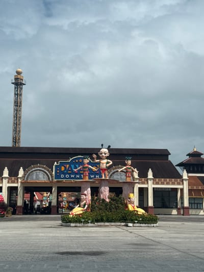 The image displays the entrance to an entertainment complex identified by a prominent sign as "DA NANG DOWNTOWN" located in Hòa Cường Ward, Vietnam. The main structure is a multi-story building featuring a dark brown, multi-pitched roof and lighter-colored facades with several arched entryways. Directly in front of the entrance, a central landscaped island features multiple colorful, stylized statues. The tallest statue depicts a light-skinned, cartoon-like figure with two antennae-like protrusions, dressed in red and green attire, with its right arm raised. It is flanked by two similar standing figures on elevated platforms and two seated figures at a lower level, all set amidst green foliage. The ground in the foreground consists of an expansive area paved with grey interlocking concrete pavers. In the background, to the far left, a tall, slender observation tower, light brown in color, with a spherical dome at its summit, rises into an overcast sky filled with varying shades of grey clouds. No human subjects or discernible activities are observed in the immediate vicinity.