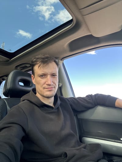 A man is seated in the driver's seat of a vehicle, looking at the camera with a neutral expression. He is wearing a black hoodie. The vehicle has a sunroof open, revealing a blue sky with scattered clouds. The interior of the car, including the dashboard and seats, is visible. The background outside the car window shows a bright sky. There is a cell tower visible in the distance through the open sunroof on the left side of the image. This image was taken in Abrykosivka, Ukraine.