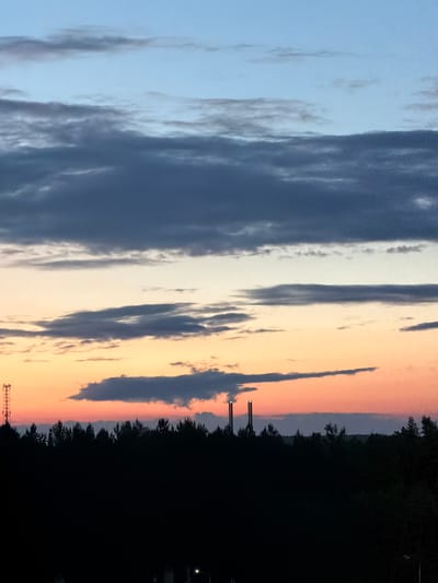 This media file depicts a sky at dusk with dramatic cloud formations. The sky transitions from a pale blue on the left to shades of orange and pink on the right, characteristic of a sunset. Dark clouds are scattered across the sky, some appearing thin and wispy, while others are dense and heavy. Silhouetted against the colorful sky is a dark treeline, suggesting a natural landscape. In the foreground, two tall, slender structures, possibly industrial chimneys or communication towers, are visible. The bottom right portion of the image is almost entirely black, obscuring any details of the ground or foreground objects, but it does appear to contain what could be a few parked cars. The overall mood is serene and evocative, hinting at the end of a day. The image appears to be taken in a location with open skies and natural surroundings, possibly in or near Tsiolkovsky, Russia, given the context. No visible text is present in the image.