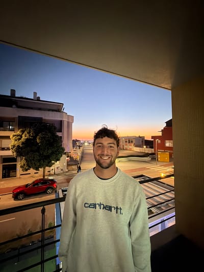 The image captures a young adult male on a balcony in Gafanha da Nazaré, Portugal, during twilight. The subject, positioned centrally, has dark curly hair, a beard, and is smiling directly at the viewer. He is wearing a light gray crew-neck sweatshirt with "carhartt." printed in dark blue across the chest.
The scene is viewed from an elevated perspective, likely a building balcony, with a dark horizontal structure overhead and a dark metal railing partially visible in the lower frame.
In the background, an urban streetscape unfolds under a sky transitioning from deep blue at the zenith to an orange and pink hue near the horizon, indicative of sunset. A bright celestial body is visible in the upper left sky.
To the viewer's left, a multi-story building with light-colored facades and darker architectural elements stands prominent. A large, densely foliated tree, illuminated by artificial light, is positioned adjacent to this building. A red crossover SUV is parked on the street below.
To the viewer's right, the street continues, revealing additional buildings, some featuring orange or red facades. Streetlights are illuminated, casting light onto the pavement. A pedestrian is visible walking on the sidewalk in the mid-ground. The overall environment is illuminated by a blend of natural twilight and various artificial light sources from the urban setting.