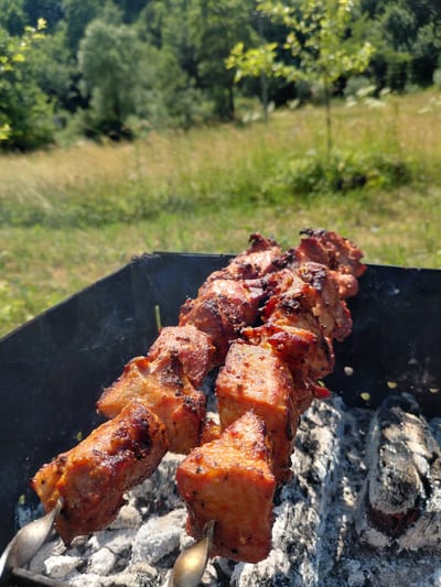 Two skewers of marinated meat are being grilled over glowing coals in a black metal grill. The meat is browned and appears to be seasoned with herbs and spices. Smoke rises from the grill, indicating that the meat is actively cooking. The grill is set up outdoors in a grassy area with trees in the background. The overall scene suggests a casual outdoor barbecue. The location is specified as Chernyk, Ukraine.