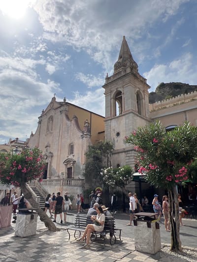 The image depicts a street scene in Taormina, Italy, with the Church of San Giuseppe and its bell tower prominently featured. The church, a historic building with a light pink facade, is partially visible on the left, while the bell tower, constructed of stone with arched openings, stands to the right of the church. In the foreground, a paved piazza is populated with various people.

Several individuals are seen walking through the piazza, some appearing to be tourists. On the left, a woman is seated on a dark-colored bench, holding a phone. In the background, more people are milling about, and some are seated at tables outside a establishment labeled "DOLCE & GABBANA". Blooming pink oleander trees add vibrant color to the scene, framing the piazza and the historic architecture. The sky above is partly cloudy, with the sun casting strong light and shadows. The overall atmosphere suggests a bustling daytime scene in a popular Italian tourist destination.