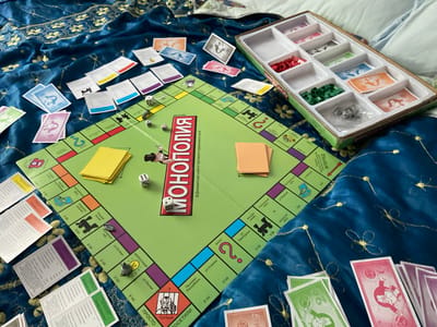A Monopoly board game is set up on a dark blue, textured fabric, possibly a blanket or tablecloth, with gold floral embroidery. The board is predominantly green with various colored property squares and text. Several cards are scattered around the board, including property deeds and chance cards, with some featuring illustrations. Dice are visible on the board, one showing a six and another showing a three. In the top right corner, a white plastic tray holds colored tokens, including red and green pieces, and some silver game tokens. A stack of yellow paper is on the board. The image appears to be taken from a slightly elevated angle. There are no people visible in the image.