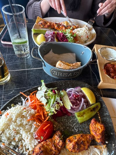 A view from above shows a table in an establishment in South Ockendon, United Kingdom. A woman with a brown sweater is sitting at the table, holding a fork and knife in her hands and eating from a plate containing rice, kebab, and salad. There is a silver bread bucket filled with bread and napkins, along with wooden serving containers with dips on the table. Another plate of food with chicken, rice, and salad is also present. A clear glass with a dark straw and lime slices is visible, as well as a smaller glass filled with a clear, yellow beverage. The table itself has a dark, marble-like texture.