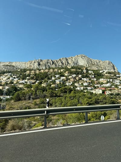 The image shows a view of the town of Calp, Spain, nestled on a hillside leading up to a large, rocky mountain. The town consists of numerous white and light-colored houses with red-tiled roofs, built closely together on the steep slope. Green trees are interspersed among the houses and cover the lower parts of the mountain. A highway guardrail runs along the bottom of the image, separating the road from the hillside. The sky is a clear, bright blue.