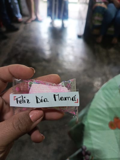 A person is holding a small, transparent plastic bag with candies inside. A white label is attached to the bag with the handwritten message "Feliz Día Mamá" (Happy Mother's Day) in black ink with red accents. The person's fingers and painted nails are visible. The background shows a blurred scene of a room with several people standing or sitting around. The floor appears to be dark and shiny. A green object is visible in the bottom right corner, along with some purplish shapes that could be grapes. The location is assumed to be Quevedo, Ecuador based on the text on the label. The scene seems to be a celebration of Mother's Day.