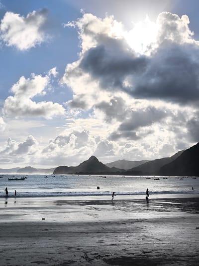 This vertical photograph depicts a coastal scene at Selongbelanak, Indonesia, characterized by dramatic natural light and a dynamic sky. The time of day appears to be late afternoon or early morning, indicated by the strong backlighting and the sun's position.

The upper half of the image is dominated by a vast sky filled with an array of clouds: bright white, fluffy cumulus clouds mingle with darker, more ominous grey formations. The sun is partially obscured by a large, luminous cloud in the upper right, casting intense light and visible rays downwards.

Below the sky, the calm ocean stretches across the mid-ground. A distant shoreline of rolling hills and mountains, largely appearing as dark silhouettes, frames the scene. Numerous small, traditional boats are scattered across the water, particularly near the base of the hills, suggesting local fishing or transportation activities.

In the foreground, a wide expanse of wet, dark sand reflects the bright sky and clouds, creating shimmering patterns. Several silhouetted figures are visible on the beach and in the shallow water. On the left, an adult and a child stand by the water's edge. Towards the center, a smaller figure runs along the sand, while another person walks into the frame on the right. These figures appear to be engaging in leisure activities, enjoying the beach.

The image is notable for its strong contrast between the bright sky and the dark silhouettes of the land, boats, and people, enhancing the atmospheric quality. No visible text is present in the media file.