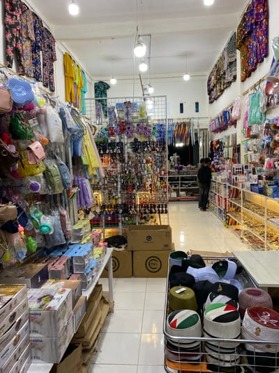The image shows the interior of a small store in Lamteumen Barat, Indonesia, filled with a variety of merchandise. The store is brightly lit by overhead fluorescent lights. Racks and shelves are densely packed with items, including clothing, accessories, and packaged goods.
On the left side of the image, hanging clothing and accessories are displayed, such as floral patterned pajamas, colorful tops, handbags, and smaller items like keychains and decorative objects. Further back, there are racks of what appear to be umbrellas and more hanging items.
In the center of the store, a large wire rack contains an assortment of hats, including skullcaps and kufi hats in various colors and designs. Some of these hats are decorated with national flags, such as the Palestinian flag. Adjacent to the hat display are boxes of packaged goods, possibly kitchenware or home goods, stacked on shelves and a table.
On the right side of the store, shelves display more merchandise, including what look like electronic accessories or phone cases, as well as more general retail items. Further back, there is a glimpse of another section of the store with more shelving.
Two individuals, appearing to be men, are standing in the middle-right section of the store, looking at the merchandise on the shelves. One is wearing a black long-sleeved shirt and dark pants, and the other is wearing a dark olive green shirt and dark pants. They appear to be customers or employees.
The flooring is tiled, and the walls are painted a light color. The overall impression is of a busy, well-stocked retail space.