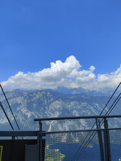 The image depicts a scenic view from a high vantage point, likely a viewing platform or cable car, overlooking a vast mountain range under a clear blue sky. The foreground features a metal railing with a safety net, suggesting an observation area. Cable car wires are visible on either side of the frame, angled downwards, reinforcing the idea of being elevated.
Beyond the railing, a breathtaking panorama of rugged mountains unfolds. The mountains are covered with dense green forests on their lower slopes, transitioning to rocky, barren terrain higher up. The scale of the mountains is immense, with numerous peaks stretching towards the horizon. A large, fluffy white cloud is prominently positioned in the sky, creating a striking contrast with the deep blue. The lighting suggests it is daytime, with the sun casting clear shadows and highlighting the textures of the landscape. The overall mood is one of awe and natural beauty.
The scene is likely located in Ukanc, Slovenia, given the mountainous terrain and potential for cable car access to such viewpoints. The presence of a body of water, possibly a lake, is discernible at the very bottom of the image, peeking through the netting of the railing. No people are visible in the image.