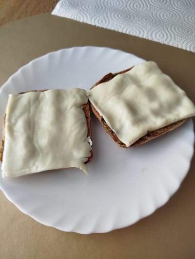 Two pieces of dark bread, each topped with a slice of white cheese, sit on a white plate. The plate is positioned on a brown surface. A white paper towel with a circular pattern is visible in the upper right corner of the image. The scene is indoors, likely in a domestic setting. There are no people or animals present. The lighting suggests it is daytime, possibly natural light from a window. The focus is on the food, with the background slightly blurred. The overall impression is of a simple snack or meal preparation.