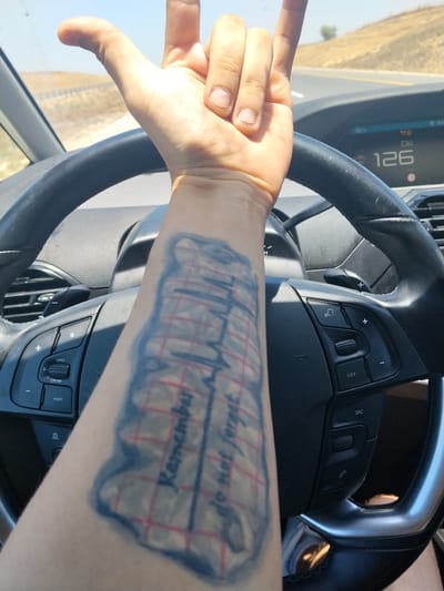 A close-up shot shows a person's left arm and hand, adorned with a tattoo, resting on a car's steering wheel. The tattoo, visible on the forearm, features a red-lined grid pattern overlaid with text that reads "Remember do not forget." The hand is positioned with the thumb and index finger extended upwards, forming a "rock on" gesture, while the middle, ring, and pinky fingers are curled down. The steering wheel has several buttons on its left side, including "LIMIT," "0-CRUISE," and "MEM." The dashboard displays a speed of "126." The scene appears to be from inside a car traveling on a road, with a dry, hilly landscape visible through the windshield. The lighting suggests it is daytime. The location is specified as Yoav Regional Council, Israel.