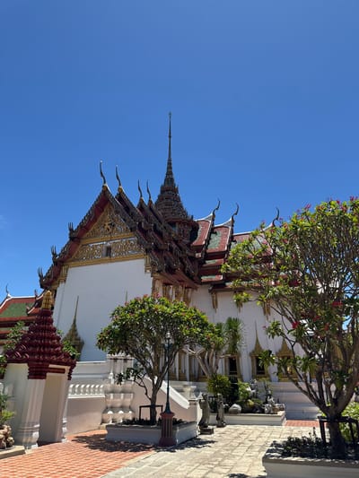 The image shows a building in Bang Pu Subdistrict Municipality, Thailand, under a clear blue sky. The building has a white facade with ornate gold details and a multi-tiered roof with red, green, and gold accents. Decorative pointed finials extend from the roof edges. A tall, tapering spire rises from the top of the roof. Trees with green leaves and red flowers are positioned near the building. A portion of a brick-paved area is visible in the foreground. The overall composition suggests a traditional Thai architectural style.