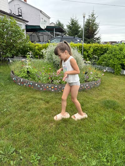 A young girl with a ponytail, wearing a white tank top and grey shorts, stands in a grassy backyard. She is wearing light-colored slides on her feet. The girl is looking down and appears to be reaching towards a circular flower bed in front of her. The flower bed is edged with a concrete border decorated with colorful mosaic tiles. Behind the flower bed, a variety of green bushes and trees are visible. In the background, a house with a satellite dish on the roof and a carport with a vehicle parked under it can be seen. The sky is overcast. The setting appears to be a suburban residential area in the city of Michurinskoye, Russia.