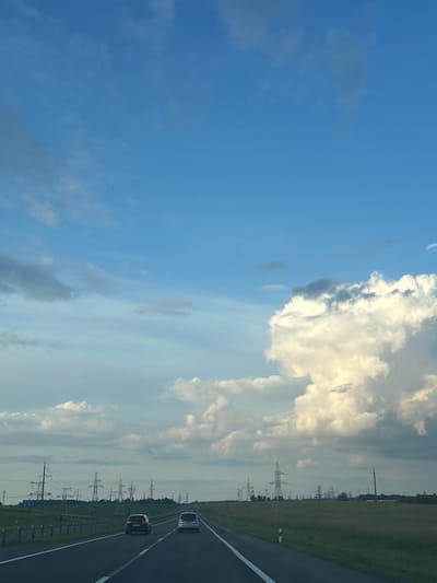 The image captures a view from a vehicle traveling on a highway in the Старасвержанскі сельскі Савет, Belarus. The sky is a vibrant blue with scattered white and grey clouds. On the right side of the frame, a large, illuminated cumulus cloud dominates the upper portion of the sky. The highway is paved with asphalt and marked with white lines. Two cars are visible on the highway, with a dark-colored car in the foreground and a lighter-colored car ahead. The landscape flanking the highway is characterized by green fields and numerous power line towers stretching across the horizon. A road sign with the number "283" is visible on the left side of the highway. The overall scene depicts a typical rural highway landscape in Belarus under a partly cloudy sky.