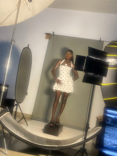A fashion photoshoot is taking place in an indoor studio in South Ropp, Nigeria. A young Black woman is posing in the center of the frame, wearing a short, white, polka-dot dress and black sandals. She has her hand on her hip and her gaze is directed towards the camera. The studio is equipped with professional lighting equipment, including large softboxes and flashes, as well as a backdrop. A monitor displaying an image is visible in the upper right corner of the frame. The overall atmosphere is professional and creative.