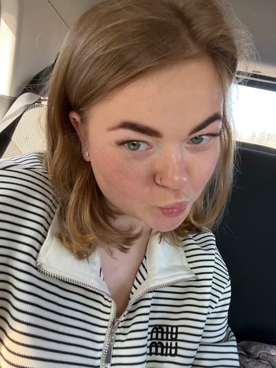 The image is a close-up selfie of a young woman with light brown hair and green eyes. She has distinct dark eyebrows and a silver hoop septum piercing. Her lips are slightly parted and puckered, conveying a playful or pouting expression. On her visible earlobe, there are three stud piercings, decreasing in size.

She is wearing a white full-zip jacket or sweater with thin black vertical stripes. The brand "MIUMIU" is embroidered in black on the right side of the garment, near the chest area. A silver-toned zipper runs down the front of the jacket.

The setting appears to be the interior of a vehicle, possibly a car, as indicated by a visible seatbelt strap on the left and the ambient light suggesting a window. The bright, natural illumination indicates it is daytime. No specific visual cues in the image directly identify the geographical location as Beloborodovo, Russia. The focus is primarily on the woman's face and upper body.
