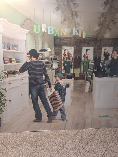 The image depicts a life-size advertisement or mural of a retail store interior, possibly for children's clothing, as suggested by the "URBAN KIDS" lettering. The scene is set within a brightly lit store with light-colored wooden floors and white shelving units filled with folded clothes and books. Several mannequins or figures are present, including a woman on the right talking on a cell phone, a man in a striped shirt and jeans looking at the shelves, and a young boy in a plaid shirt and green baseball cap holding a striped shopping bag. The background features brick walls and posters of children. The advertisement is positioned at ground level, with a granite-tiled floor in the foreground, suggesting it's located in a public area. The image does not provide any specific information about a location in Bochum, Germany.