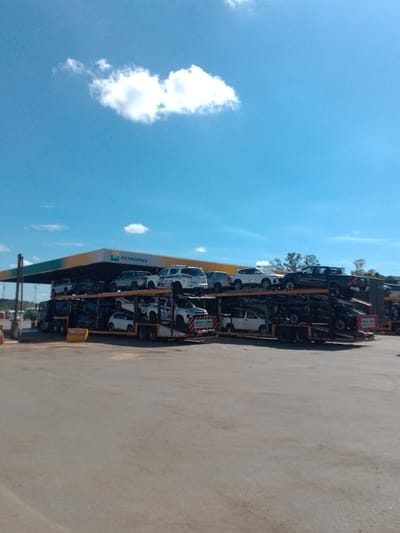An eye-level, medium shot captures a car carrier truck loaded with vehicles parked in front of a BR Petrobras gas station under a bright blue sky with scattered white clouds. The gas station canopy, with its distinctive yellow and green design, is visible in the background. The car carrier, a large multi-level trailer, is filled with a variety of cars and trucks, some of which appear to be police vehicles based on their markings. The truck is parked on a paved surface, likely a road or parking lot, in front of the gas station. The overall scene suggests a logistical operation, possibly involving the transport or delivery of vehicles to the gas station or a nearby location in Cáceres, Brazil.