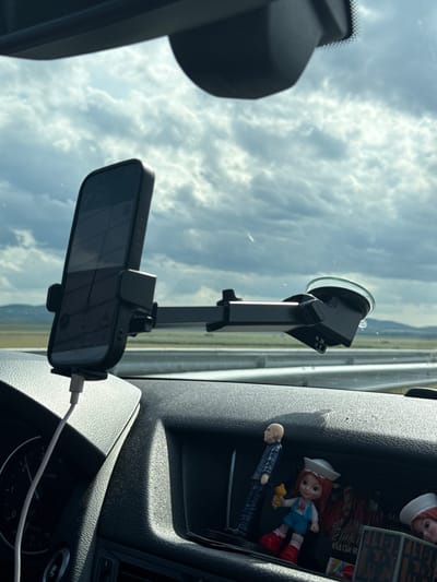 A smartphone is mounted on a windshield by a suction cup mount, with the phone displaying a navigation app. The car's dashboard is visible below the mount, featuring a charging cable plugged into the phone. Several small figurines, including a bald man in a suit and a girl in a sailor outfit holding an ice cream cone, are placed on the dashboard. The car is on a road, indicated by the presence of a highway barrier and a landscape of fields and hills under a cloudy sky. The scene is set in the city of Polsko Padarevo, Bulgaria.