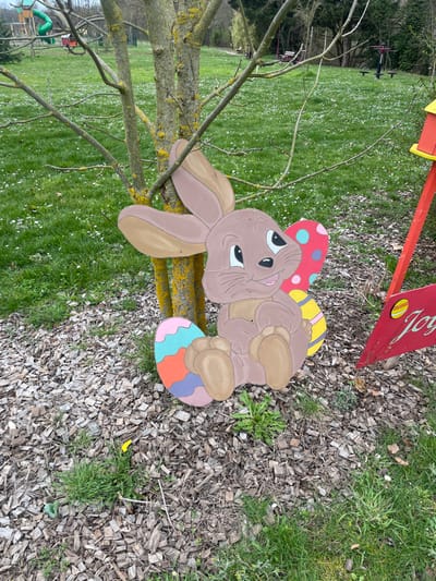 The image displays an outdoor scene in a park or recreational area in Holtzheim, France. In the foreground, a brown-painted wooden Easter bunny decoration is positioned against the base of a multi-trunked tree with yellow-green lichen on its bark. The bunny figure is depicted smiling, holding a pink Easter egg with blue and white polka dots. A second striped Easter egg, colored blue, orange, and purple, rests to the bunny's left, and a third yellow striped egg is partially visible behind the bunny on the right.

The immediate ground surrounding the bunny is covered with light brown wood chips, with sparse green grass and weeds growing through them. Beyond this, a wide expanse of green lawn, dotted with numerous small white flowers, stretches into the mid-ground. In the background, various playground structures are visible, including a prominent green slide, a red structure, and other brown wooden play elements. Bare-branched trees and some evergreen foliage line the distant perimeter of the area. To the right of the bunny, a partially visible red wooden structure, possibly a birdhouse or decorative element, stands on a red post, with a red banner or sign featuring yellow accents beneath it, part of the word "Joy" discernible. No individuals are present in the scene.