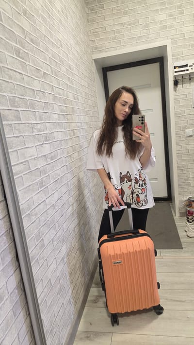 A woman with long, dark, wavy hair takes a selfie in a hallway with a light gray brick-patterned wallpaper. She is wearing a white t-shirt with cartoon dogs printed on it and black leggings. In her right hand, she holds a smartphone, and in her left hand, she grips the handle of a small, orange suitcase with wheels. The suitcase is positioned on the floor next to her. Behind her, a white door is visible, and to the right, a shelf with keys hangs on the wall. The floor is made of light-colored wood planks. The scene appears to be an indoor residential setting, likely a hallway or entryway in a home.