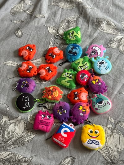 On a gray floral patterned surface are scattered 21 toy keychains with cartoon monster designs. The toy designs include monster faces, soda can shapes, and snack faces. The environment is devoid of any people or location context beyond the floral pattern background. The scene contains no indication of being taken in Chuprino, Russia.