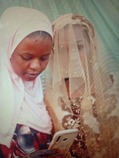 The scene, located in Sumaila, Nigeria, features two individuals in an indoor setting with a light green background. The individual on the left, displaying a light brown complexion, wears a light pink hijab and a partially visible red garment. This individual is holding a white smartphone in their right hand, which is adorned with dark henna patterns and red nail polish, and is looking downwards towards the device. The individual on the right is partially obscured by a golden-beige veil embellished with small white pearls, revealing only a faint outline of an eye. This person wears a dark red garment featuring intricate gold floral embroidery. Both individuals are in close proximity, with their gaze suggesting shared attention towards the smartphone.