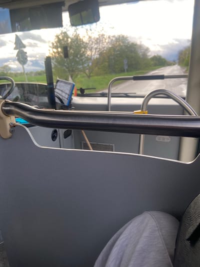 The image captures an interior view from a vehicle, likely a bus or coach, looking forward through the windshield. The perspective is from the front passenger area, slightly to the right, with a dark grey partition or wall prominent on the right side. Several polished metal grab bars or poles are visible in the foreground and midground, one extending vertically and another forming a curved handle with a yellow grip. The base of one bar is secured by two screws.
On the right side of the frame, part of a person's lap or leg, clad in a grey hooded sweatshirt or similar comfortable clothing, is visible, suggesting a passenger is seated nearby.
Through the front windshield, a bright, cloudy sky dominates the upper left, casting significant glare and reflections on the glass. Trees with green foliage are visible along what appears to be a road or pathway, suggesting an outdoor, possibly rural or semi-urban, setting. A rearview mirror is visible on the left side of the windshield, and parts of the vehicle's dashboard, including what looks like a mounted electronic device or screen, are also discernible. The overall lighting suggests it is daytime, possibly a bright, overcast day or a period with strong sunlight.
The scene takes place in a vehicle in Golyanovtsi, Bulgaria. No readable text is visible.