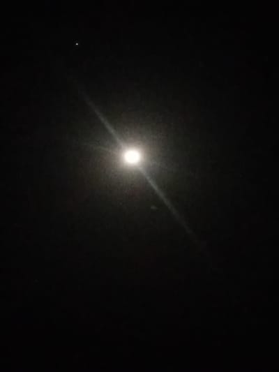 The image depicts a dark night sky over Nata-Ala, Nigeria. A prominent, bright circular light source, consistent with the moon, is situated slightly below the center, exhibiting a distinct starburst lens flare emanating diagonally upward and to the lower left. A smaller, fainter point of light, interpreted as a star or distant planet, is visible in the upper left quadrant. The environment is dark and devoid of artificial structures, ground features, or human presence. No discernible actions or interactions are present other than light emission and observation.