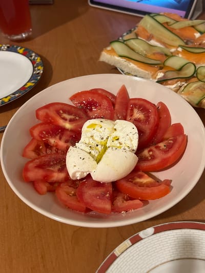 A caprese salad is served on a white plate, with sliced tomatoes and a ball of fresh mozzarella in the center, drizzled with olive oil and sprinkled with pepper and salt. The salad is arranged on a wooden table. To the right of the salad is a plate with appetizers, possibly smoked salmon and cream cheese on bread, topped with cucumber slices. In the background, there is a red drink in a glass and another plate with a decorative rim. The scene appears to be indoors, possibly in a home or restaurant setting, but no specific location in Skawina, Poland can be identified.