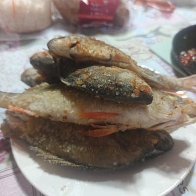 A stack of four fried fish lies on a white plate. The fish are golden brown and appear to be small, whole fish. Some of the fish show remnants of orange fins. In the background, a blurred image of a food package and a black bowl with a red substance can be seen. The setting appears to be a domestic environment, possibly a kitchen or dining area.