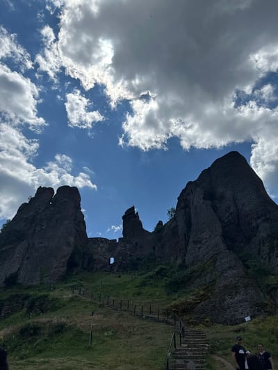 The image depicts a view of the Belogradchik Rocks, a UNESCO World Heritage Tentative List site in Belogradchik, Bulgaria. The foreground features a grassy slope with a stone staircase ascending to the right, bordered by a wooden railing. A second, less distinct wooden railing follows a path winding up the hill to the left. Several people are visible at the bottom right of the frame, appearing to be tourists.
Dominating the mid-ground and background are the massive, reddish-brown rock formations characteristic of the Belogradchik Rocks. These formations rise dramatically against a blue sky filled with white and gray clouds. Nestled among the rocks in the mid-ground, part of an ancient fortress is visible, constructed of stone blocks and featuring a small arched window. The fortress walls appear to be integrated with the natural rock formations. The overall scene conveys a sense of historical significance and natural grandeur.