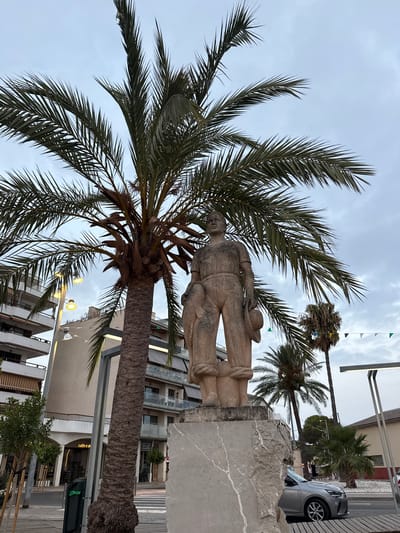 In Port d'Alcúdia, Spain, a stone statue of a man stands on a pedestal in front of a tall palm tree. The man is depicted wearing a cap and holding a net or sack. Behind the statue and palm tree, buildings with balconies are visible, suggesting an urban setting. The sky is overcast. A silver car is parked on the right side of the frame. Small flags are strung across the scene in the background.