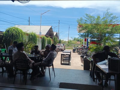 A group of men are seated at tables outside a building with a red and white canopy. The building appears to be a gas station with vehicles parked in front. Some of the men are wearing traditional clothing and headwear. The scene is set on a sunny day with a clear blue sky.