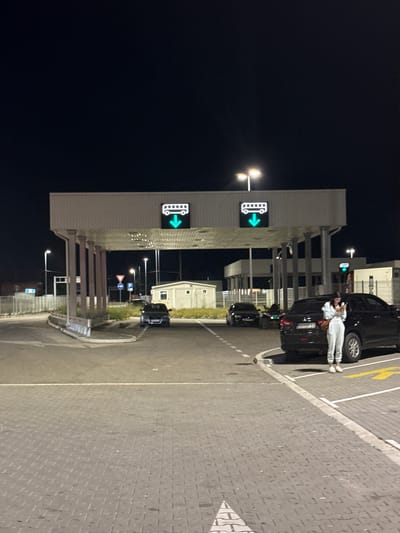 At night in Prijepolje Municipality, Serbia, a row of automated gates is illuminated with directional signage featuring a green arrow and bus icon. Several dark-colored vehicles are lined up on the right side of the gates. A person is leaning against one of the vehicles, wearing light-colored clothing. Streetlights illuminate the area, and various traffic signs are visible beyond the gates. The ground is paved with gray blocks, marked with white and yellow lines. The sky is dark.