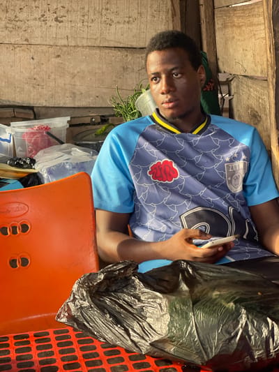 A young man with dark skin and short, dark hair is depicted in a portrait-style shot, looking directly at the camera with a neutral expression. He is wearing a light blue short-sleeved t-shirt with a dark blue overlay on the torso that features a light blue and white abstract, repeating pattern. The collar has a yellow and black striped trim.
On the left side of his chest (viewer's right), there is a prominent red, stylized patch that resembles an internal organ or brain-like structure. On his right shoulder area (viewer's left), a white rectangular patch with the text "ALLZ" and a small crest-like symbol is visible. A large white number "6" is emblazoned on the right side of the shirt's torso (viewer's left).
He is shown holding a slender, white cylindrical object, possibly a cigarette or vape pen, between the thumb and forefinger of his right hand. His right arm rests on an orange surface, which appears to be a plastic chair or table, identifiable by a textured, grid-like pattern on its right side. Next to his arm, on the orange surface, sits a black plastic bag that appears full.
The background is simple and features light-colored wooden planks or panels. To his left, behind him, some green leafy plants or vegetables are visible in a translucent white container. Other clear plastic containers are present in the lower left background. The overall setting appears informal, possibly indoors or in a covered area.