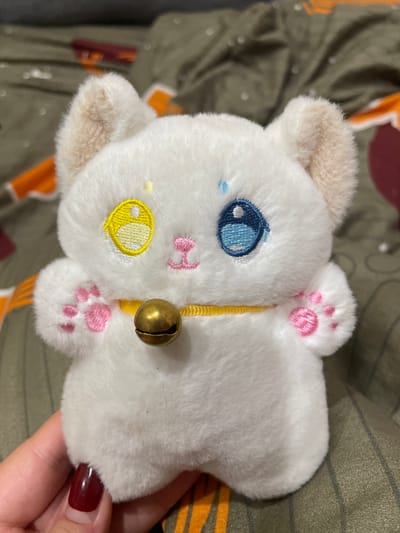 A close-up shot captures a white, fluffy stuffed toy in the shape of a cat's head, lying on a textured surface with green and orange stripes. The toy features a large blue eye with a white pupil, a small blue teardrop-shaped detail below the eye, and a pink embroidered nose. A yellow embroidered oval shape with a white circle inside is on the left side of the toy's face, resembling a cheek. Pink embroidered paw prints are visible on the top of the toy's head, and another set of pink paw prints are on the right side. A thin yellow string goes around the toy's neck, adorned with a small, brass-colored bell. A human hand with dark red nail polish is holding the toy, obscuring part of its right side. The background suggests an indoor setting.