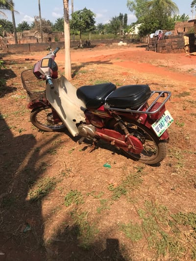 The scene, located in Adum Afo, Nigeria, depicts an outdoor area under clear daytime conditions. A red and off-white/cream colored motorcycle is parked on its kickstand on a reddish-brown dirt surface, angled slightly right of center. The motorcycle features a black mesh front basket, two distinct black padded seats, and a silver-colored metal luggage rack over the rear fender. A green and white Nigerian license plate, reading "IKOBT OF 047-KPA," is affixed to the rear.
The ground consists of dry, reddish-brown soil, sparsely covered with patches of small green leafy plants and dry grass. A small piece of green plastic debris is visible on the ground near the motorcycle's rear wheel. Distinct shadows, including that of the motorcycle, indicate strong overhead sunlight.
In the background, a tall, slender palm tree trunk is visible behind the motorcycle. Beyond the immediate foreground, the terrain is also reddish-brown dirt with scattered green trees, including additional palm trees. To the right, a large stack of reddish-brown bricks is present alongside a partial, unfinished building structure. Laundry is visible hanging on a line or structure in the far background to the right. No individuals are present in the image.