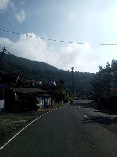 A street scene in a mountainous region under a bright, partly cloudy sky. The road, asphalt, curves gently uphill and is marked with white lane lines. A tower stands amidst trees and buildings. Motorbikes travel along the road. On the left, buildings with a sign that reads "VILLA KAMAR + KOLAM RENANG". Power lines extend across the upper part of the frame.