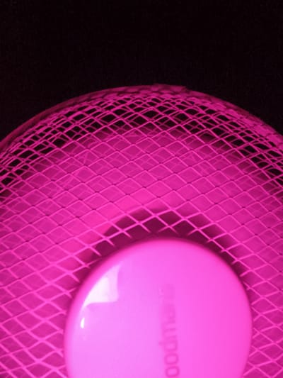 The image displays a close-up, low-angle, abstract shot of the pink grille of a fan, with a portion of the fan's motor housing visible in the lower center. The background is entirely black, suggesting a dark environment. The fan grille is a diamond-shaped mesh with a distinct pink hue. The motor housing is a smooth, rounded, lighter pink object with some indistinct text visible, possibly a brand name. The scene offers no contextual information about its location in Seliște, Moldova, or any details about people, actions, or interactions. The image's composition is focused on texture and color, making it a stylized representation of a common household object.
