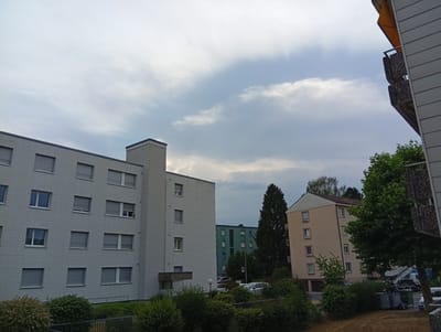 This is a daytime, overcast photograph of a residential area with apartment buildings. In the foreground, the left side of a modern, light-grey apartment building with numerous windows is visible. To the right, a tan-colored apartment building with a red-tiled roof is seen, also with several windows. Between these buildings, a taller, dark green building with many small windows is partially visible. Several trees are scattered throughout the scene, with a large green tree in the right foreground. In the background, there are other apartment buildings and some parked cars. The sky is filled with clouds. The overall impression is one of a slightly gloomy, ordinary day in a suburban or urban residential neighborhood. There is no visible text in the image.