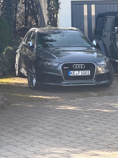 The image displays an outdoor scene in Kempten (Allgäu), Germany, under bright daylight. A dark grey Audi RS 6 Avant, bearing the German license plate KE·T 1001, is parked facing the viewer. The vehicle's front grille, featuring the RS 6 badge and Audi rings logo, is clearly visible, as are its alloy wheels. To the right and partially behind the Audi, a black Jeep Wrangler is visible, showing part of its front and side. Both vehicles are situated on a driveway composed of light grey interlocking paver stones, which exhibit distinct shadows, suggesting overhead sunlight. To the left of the Audi, a strip of green grass borders dense, dark green foliage, behind which bare tree branches and a white building wall are partially discernible. To the right of the Audi, a modern building facade or garage door with vertical dark grey panels is visible. No individuals are present, and no actions or interactions are depicted within the frame.