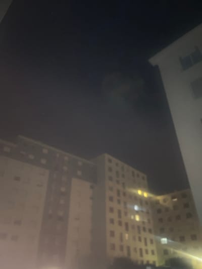 A low-light, blurry photograph captures a nighttime urban scene in a residential district of Clermont-Ferrand, France. The primary subjects are multiple multi-story apartment buildings with light-colored facades and numerous rectangular windows. One building is partially visible on the far right, while a larger complex occupies the left and center of the frame, appearing interconnected. Scattered windows within these structures are illuminated with yellow, orange, or white light, indicating interior lighting. The sky above is dark, consistent with night, displaying a faint lens flare or atmospheric haze in the upper center-right portion of the image. No people, discernible objects, or interactions are visible on the ground level or within the visible portions of the buildings. The environment is an urban residential area.