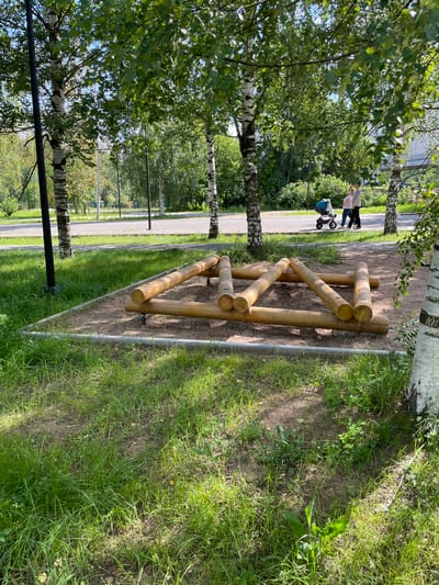 In Kirovo-Chepetsk, Russia, an outdoor fitness obstacle made of wooden logs is situated on a sand-covered area surrounded by grass and bordered by a concrete curb. The obstacle is a triangular structure formed by several logs. In the background, two people are walking on a paved path next to a street. One person is pushing a stroller. The scene is framed by birch trees with dappled sunlight filtering through their leaves.