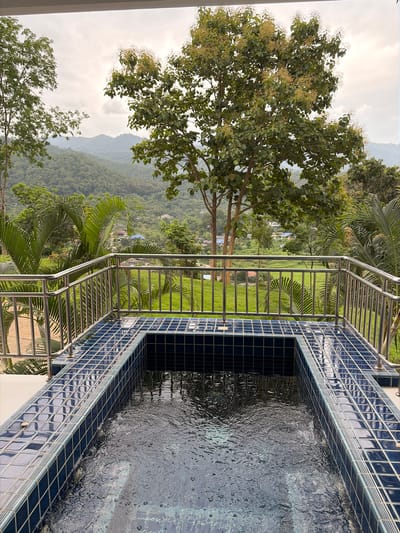 The image depicts a plunge pool with dark blue tiled edges situated on a balcony or deck. Beyond the pool, a metal railing with vertical spindles separates the foreground from a lush green landscape. The background features rolling hills covered in dense vegetation, with scattered buildings visible in the distance. Tall trees, including one with a flowering canopy, frame the view. The sky is overcast, suggesting a cloudy day. The location is identified as Ban Kao Dua, Thailand, and the scene appears to be a serene, natural setting.
