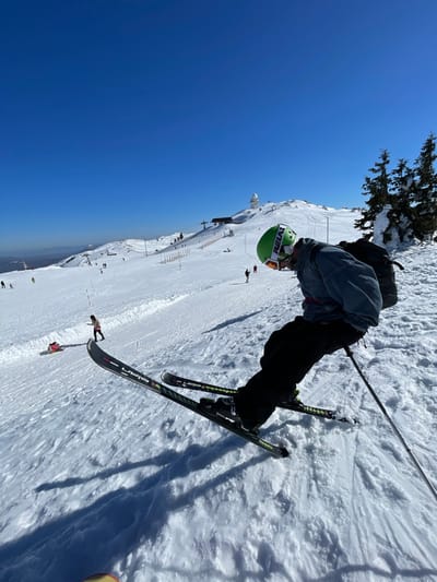 The image captures a panoramic view of a snow-covered ski resort on a clear, sunny day, identified as Jahorina, Bosnia and Herzegovina. The foreground features a skier, viewed from a low angle, actively descending a slope. This individual wears a lime green helmet with "RELAX" branding, ski goggles, a grey-blue jacket with a red stripe, black ski pants, and a black backpack. The black skis display "ELAN" branding and green accents, angled as if executing a turn. A ski pole is visible in the skier's right hand.
In the midground, the wide ski slope extends upwards and to the left, populated by multiple other individuals. To the left, a person in light clothing stands adjacent to a red sled, which appears to have a small occupant or object. Additional skiers and snowboarders are scattered across the expansive, groomed snowfield. Orange safety poles mark sections of the slope.
The background showcases the snow-covered mountain peak of Jahorina. A prominent white spherical structure, consistent with a radar dome or observatory, stands at the summit. Ski lift towers and cables are visible ascending towards this peak. A cluster of snow-laden evergreen trees is situated on the right side of the mountain. Distant mountain ranges are faintly visible on the horizon to the far left under a cloudless, vibrant blue sky.