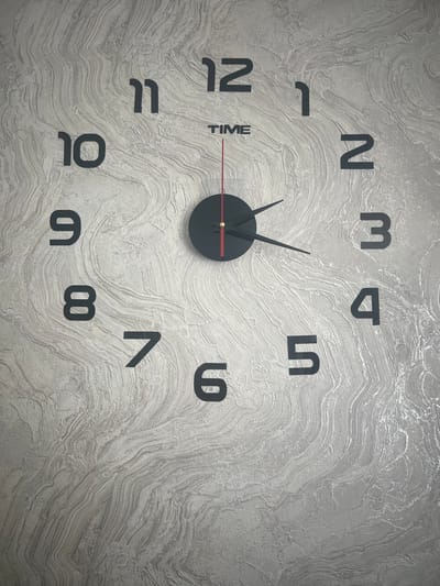 This image shows a close-up of a modern wall clock with black numbers on a textured, light-colored wall. The clock has a black circular center and black hour and minute hands, with a thin red second hand. The numbers are positioned around the clock face, indicating the hours from 1 to 12. The wall has a textured pattern that resembles wood grain or flowing water. There are no people or other objects visible in the image. The location is identified as Почтовское сельское поселение, Ukraine, though the image itself does not provide any visual cues to confirm this specific location; it is a generic indoor scene of a clock on a wall.