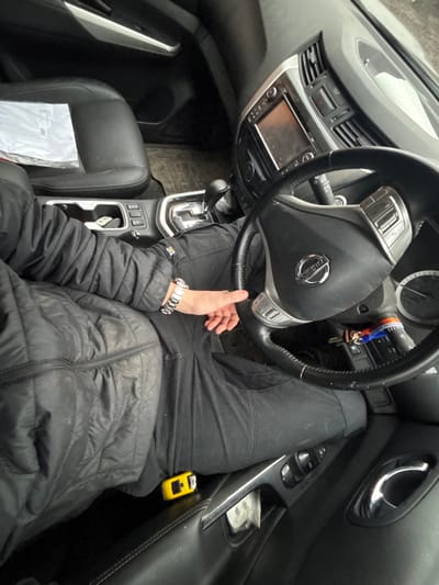 The image captures an overhead view of the interior of a car, focusing on the driver's position. A person is seated in the driver's seat, wearing a black puffer jacket and black pants. Their torso, arms, and upper legs are visible. Their left hand rests on their left thigh, near the steering wheel, and they are wearing a silver watch with a metallic strap on their left wrist. A "CAT" (Caterpillar) logo is visible on the cuff of their jacket sleeve.
The steering wheel, featuring a prominent "NISSAN" logo in the center, is black with silver accents and integrated controls. Keys with a colorful keychain are seen hanging from the ignition area behind the steering wheel. The dashboard is dark, featuring air vents, a hazard light button, and a central infotainment screen that appears dark or off. Below the screen is the gear shifter for an automatic transmission, surrounded by a chrome-like trim, and further down, cup holders and other console buttons.
To the left, part of the front passenger seat is visible, covered with what appears to be a white plastic bag or protective wrapping. The floor mat beneath the driver's footwell is dark. To the right, the driver's side door panel is visible, featuring a chrome door handle, window controls, and a storage pocket. Within this pocket, a small, distinctive yellow device, possibly a charger or tool, is placed.
The lighting suggests it is daytime, with soft, diffused light entering the vehicle. The overall impression is a static scene of someone resting in a parked car.